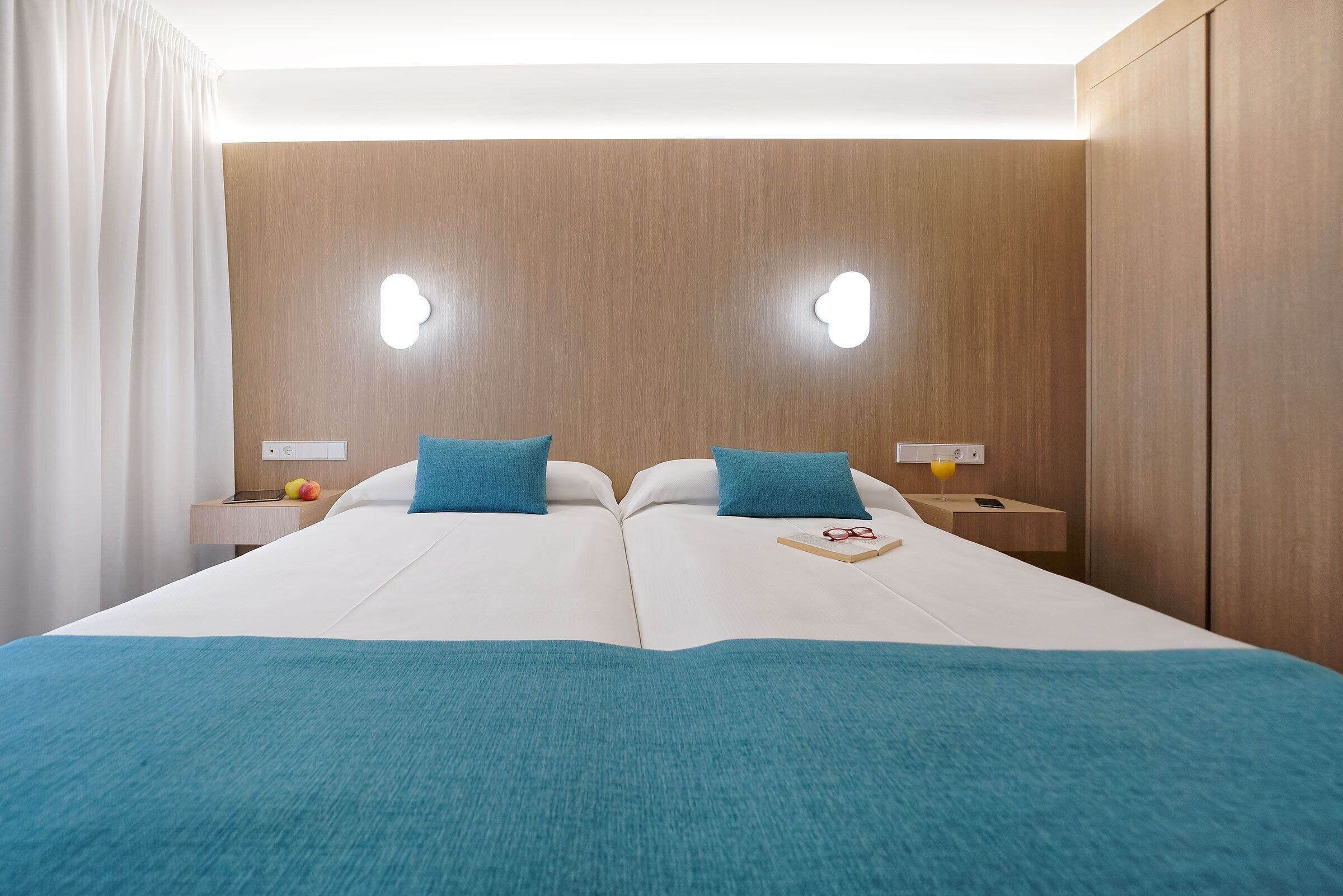 A double bed with a teal blanket, two pillows, a bedside table with a book and glasses, and a fruit bowl.