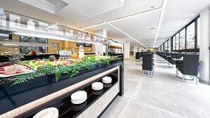 Un moderno vestíbulo de hotel con un área de bufé que ofrece diversas opciones de comida y un espacio de descanso con mesas y sillas.