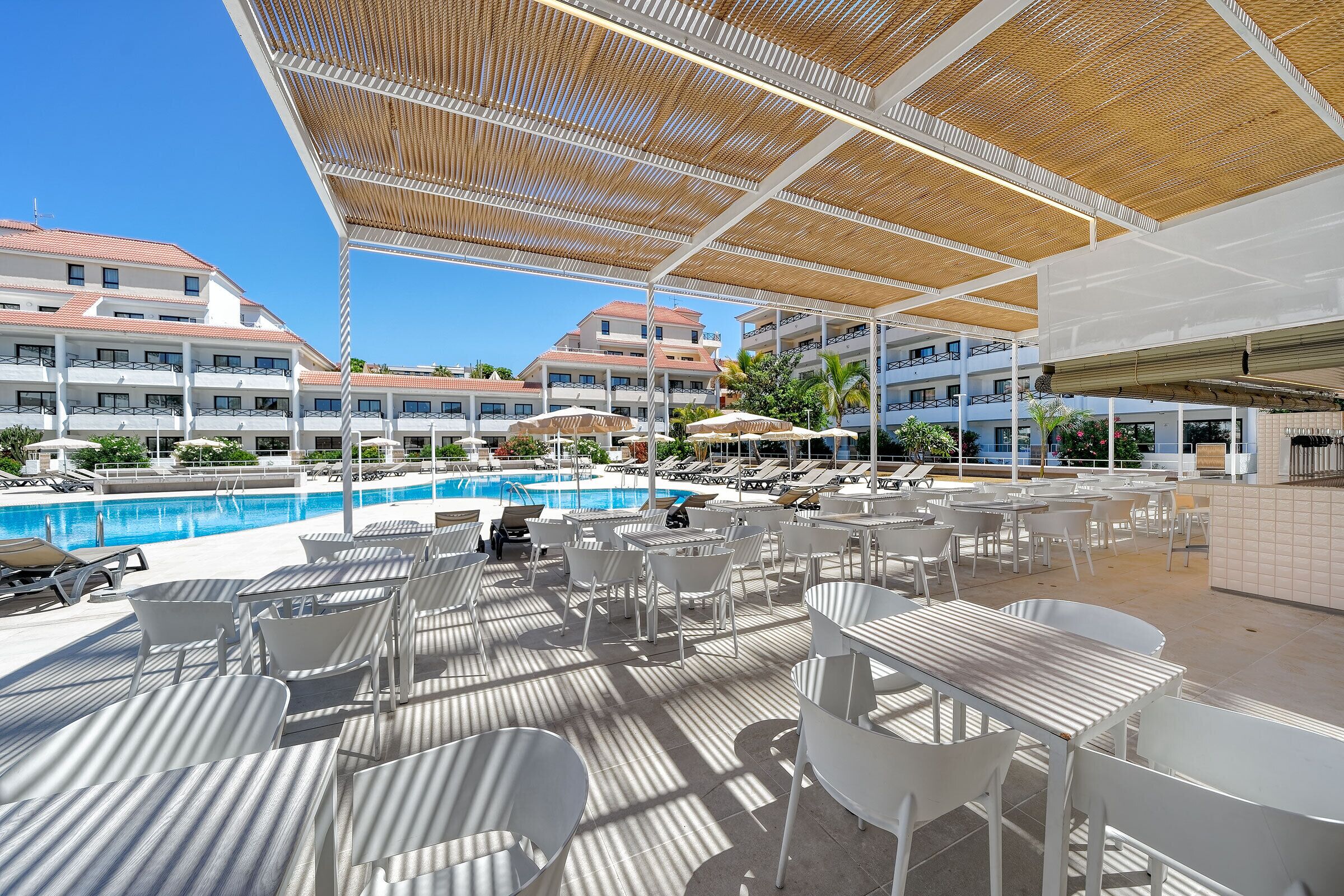 A poolside area with white chairs and tables, umbrellas, and a covered seating area with a wooden ceiling.