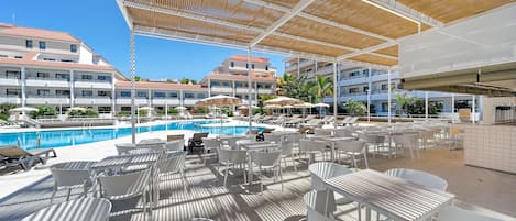 A poolside area with white chairs and tables, umbrellas, and a covered seating area with a wooden ceiling.
