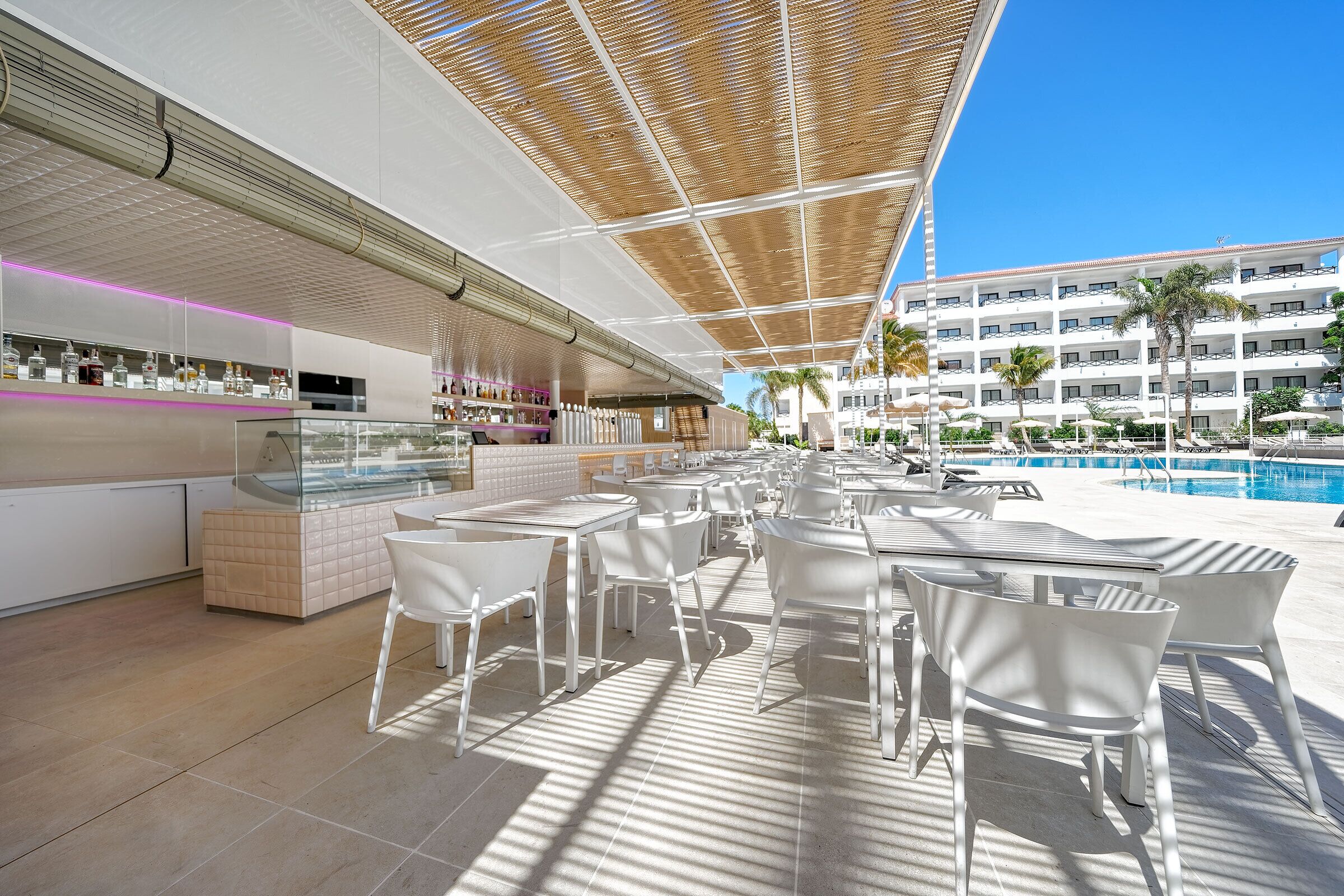 A modern outdoor bar area with white chairs and tables, a pool in the background, and a building with multiple balconies.