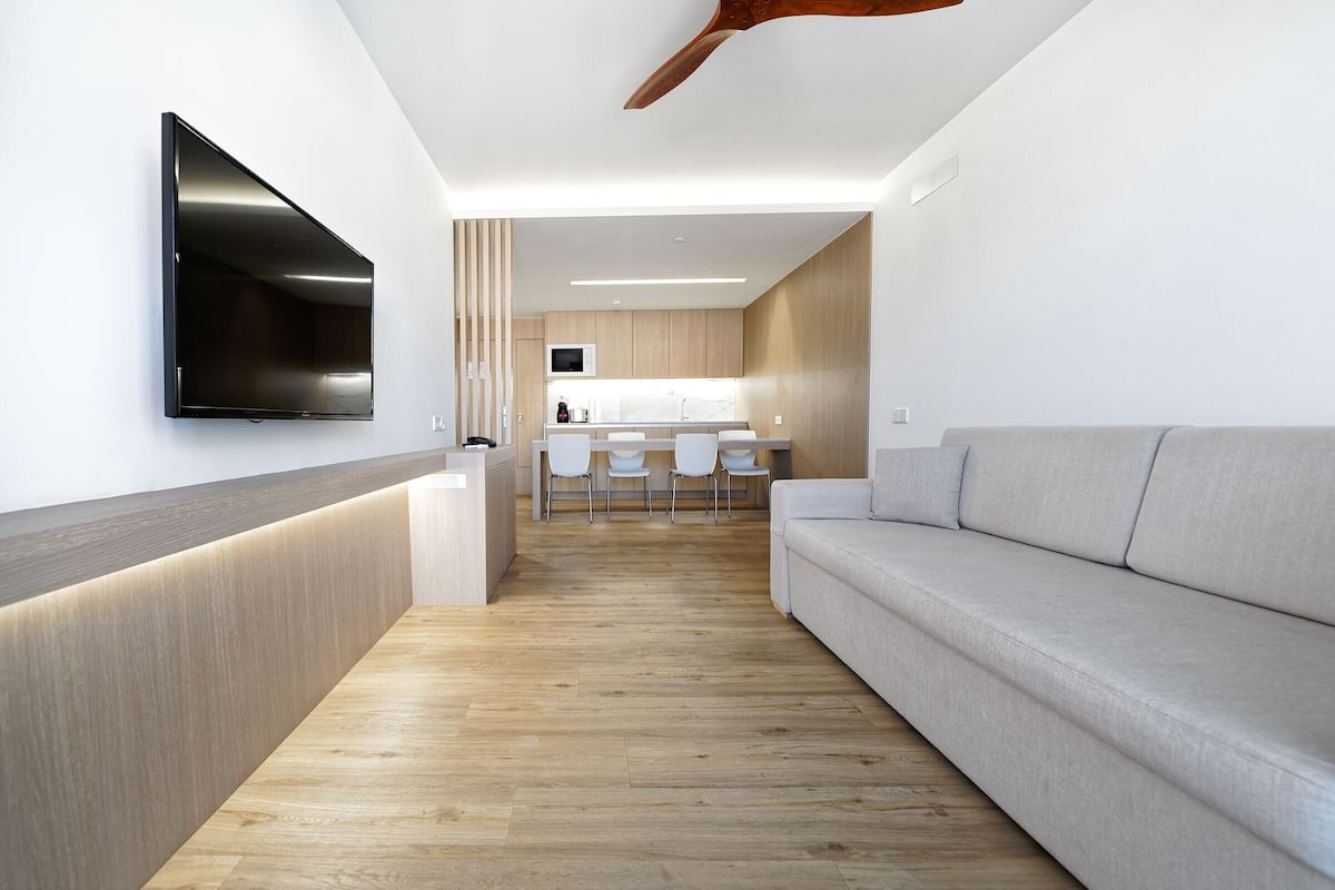 A modern living room with a flat-screen TV mounted on the wall, a wooden reception desk, a grey sofa, and a kitchen area in the background.
