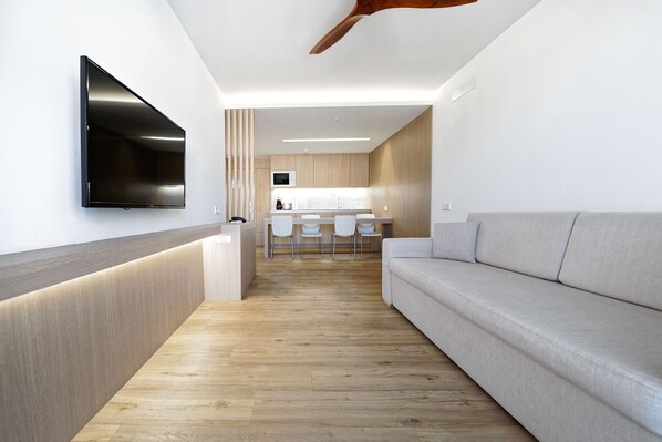 A modern living room with a flat-screen TV mounted on the wall, a wooden reception desk, a grey sofa, and a kitchen area in the background.