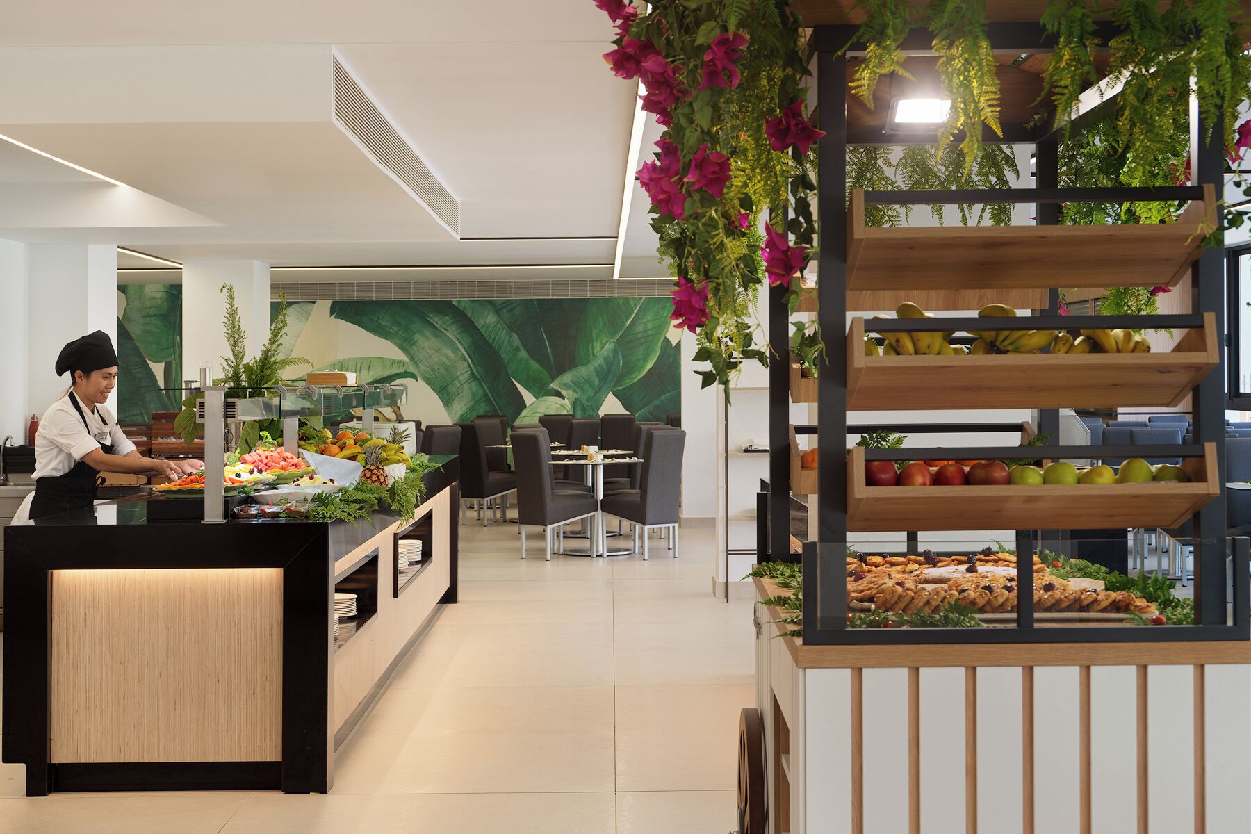 Een modern café met een kok die eten bereidt, een presentatie van fruit en een vitrine met desserts.