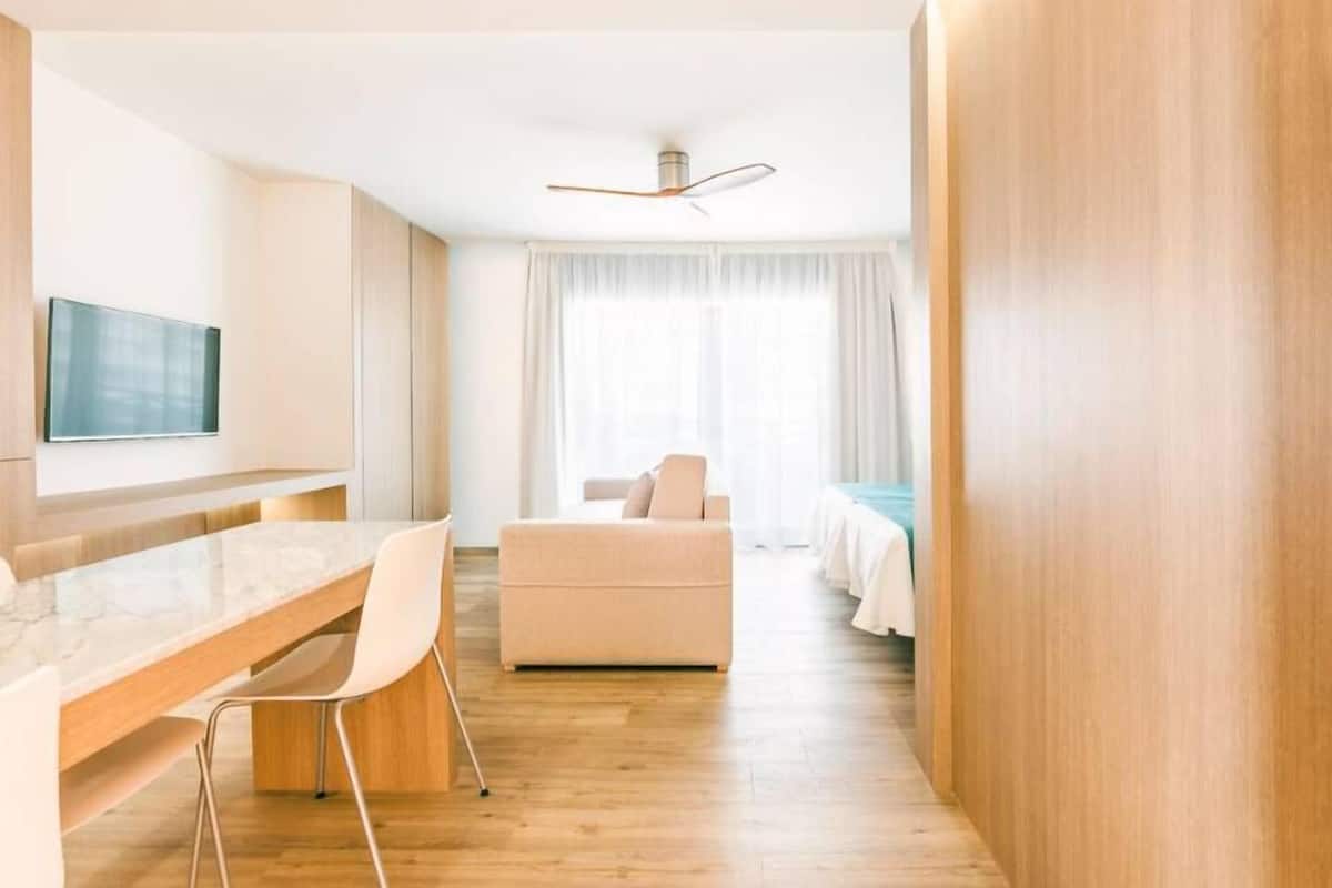 A modern hotel room with a wooden floor, a flat-screen TV mounted on the wall, a dining area with a marble countertop and two chairs, a sofa, and a bed with a blue bedspread.