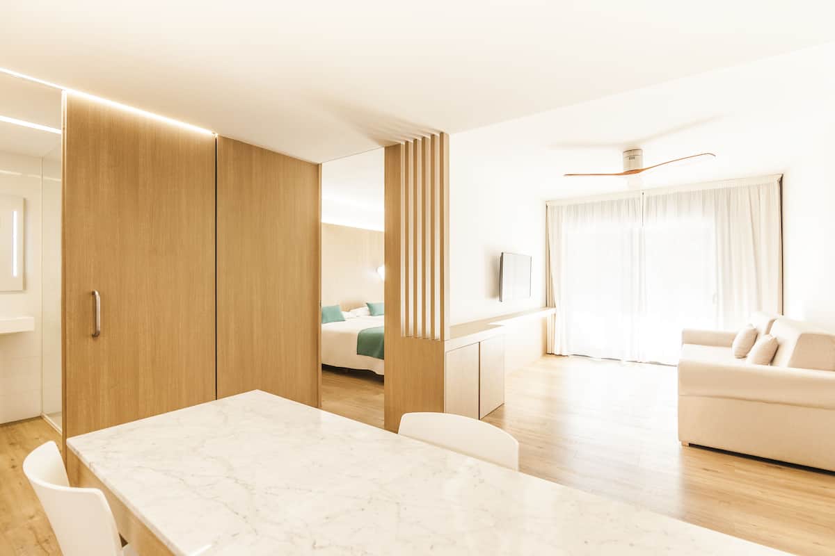 A modern hotel room with a marble dining table, a sofa, a bed with a green pillow, and a TV on the wall.
