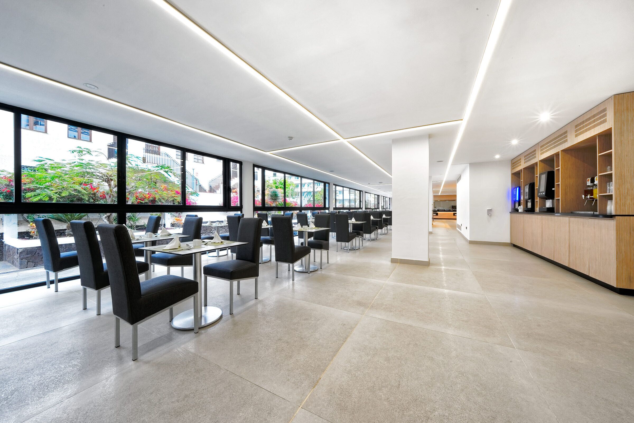 A modern dining area with long tables and chairs, large windows offering views of greenery, and a kitchen area with wooden cabinets and appliances.