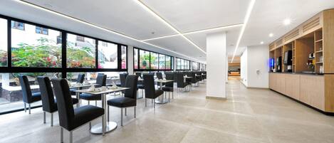 A modern dining area with long tables and chairs, large windows offering views of greenery, and a kitchen area with wooden cabinets and appliances.