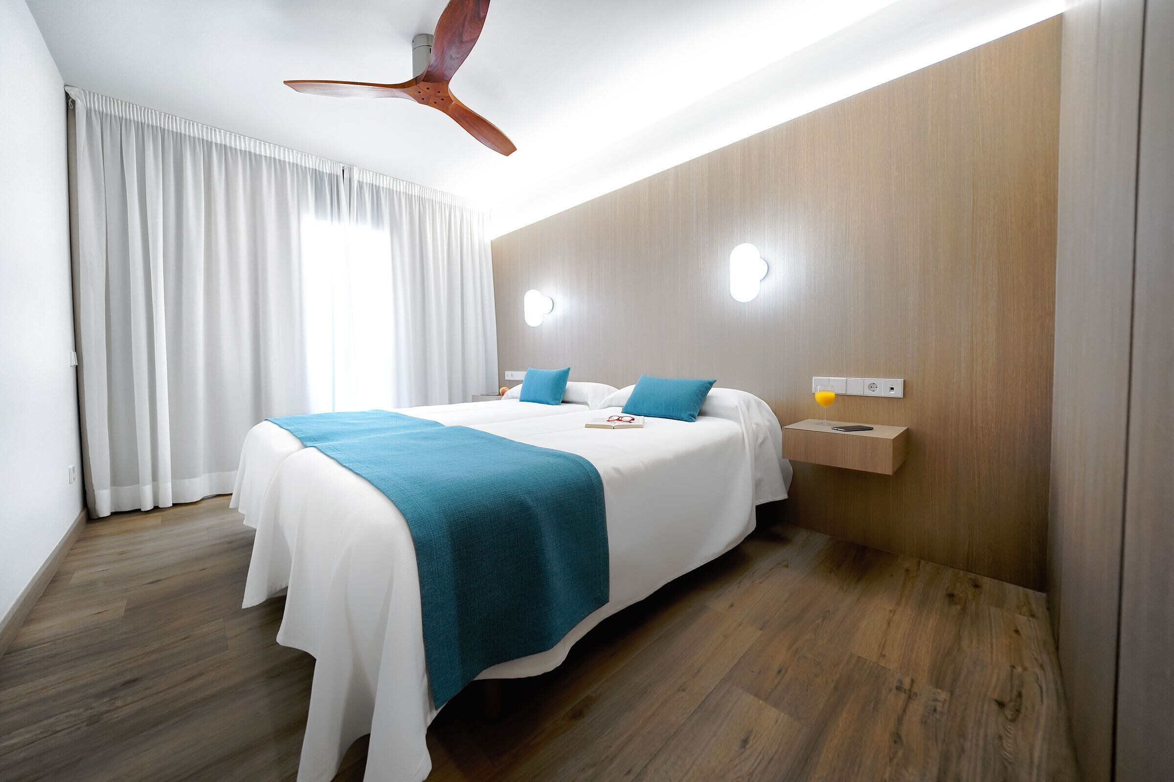 Une chambre d’hôtel moderne avec un grand lit, un ventilateur de plafond en bois, une table de chevet avec une lampe et une fenêtre avec des rideaux.