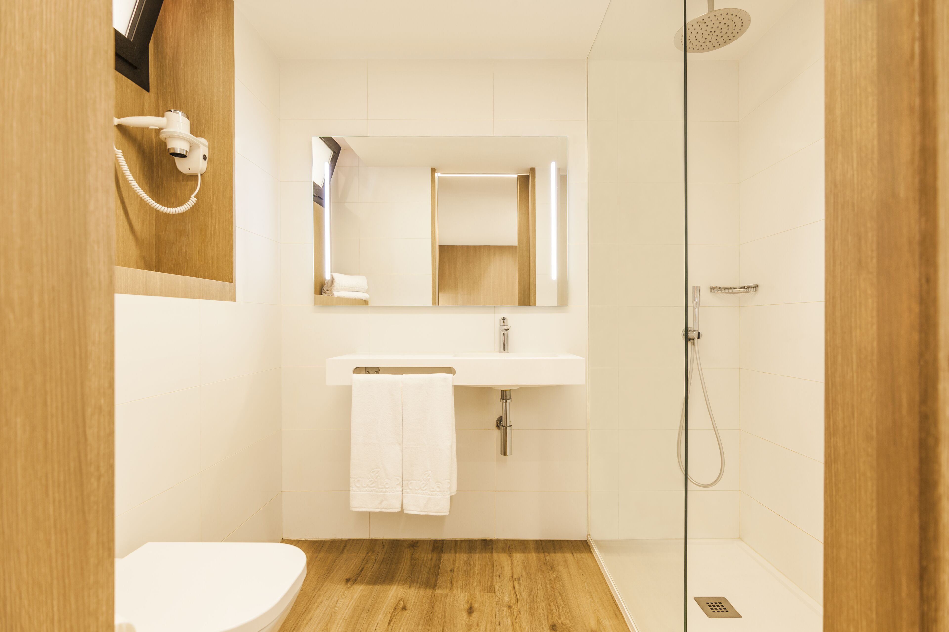 Uma casa de banho moderna com soalho em madeira, equipamentos brancos, uma pia com espelho e um chuveiro.