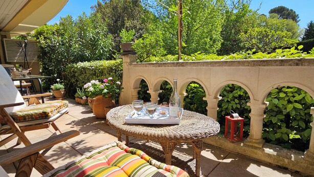Eine Terrassenfläche mit einem runden Korbstuhl-Tisch, gedeckt mit Gläsern und einer Flasche, umgeben von Sitzgelegenheiten mit Kissen und Topfpflanzen.