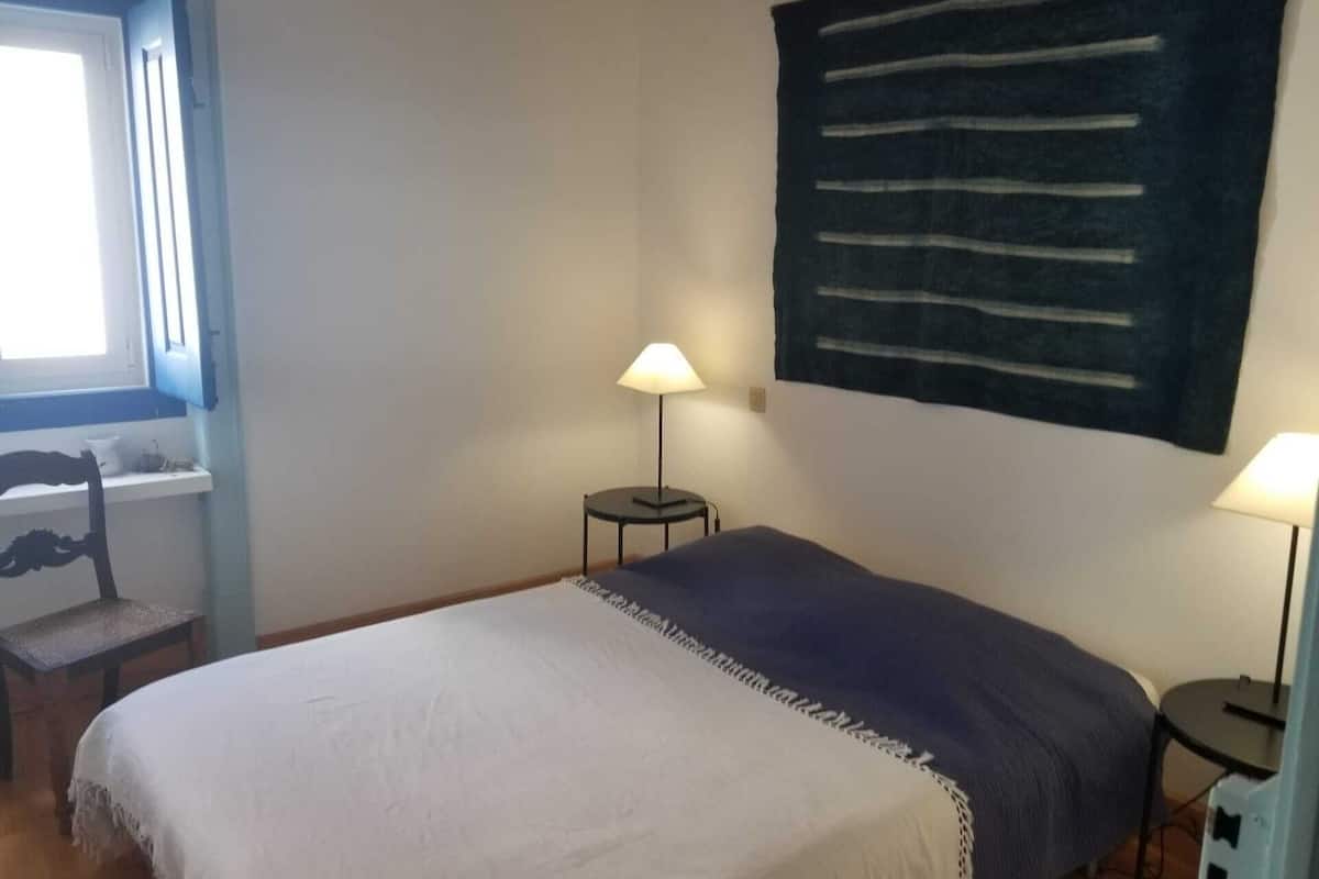 A bedroom with a bed, two bedside tables with lamps, a chair, a window, and a wall hanging.