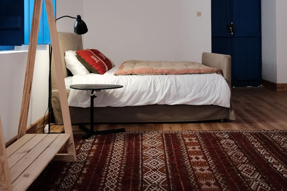 A bedroom with a bed, a wooden chair, a desk, a patterned rug, a blue wardrobe, and a hanging light fixture.