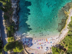 Une vue aérienne d’une plage aux eaux turquoise et limpides, avec un rivage sablonneux et des personnes se prélassant au soleil.