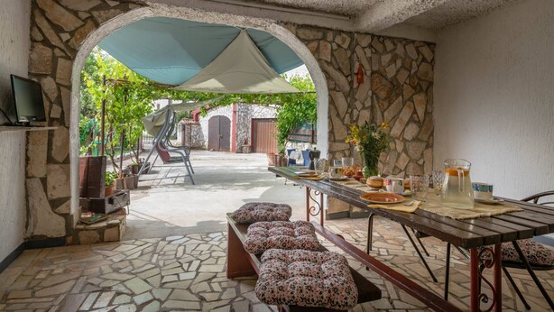 Une terrasse dallée de pierres, avec une table dressée pour le repas, des sièges d’extérieur et un téléviseur placé sur le côté gauche.