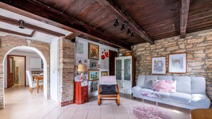 Un salon chaleureux avec un mur en pierre, un plafond en bois, un canapé gris, une chaise en bois et une petite table surmontée d’un tapis rose.