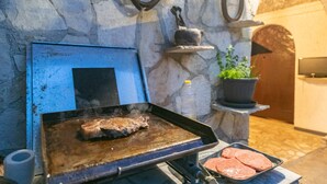 Un barbecue avec un steak en train de cuire, un plateau de viande, une bouteille d’eau et une plante en pot.