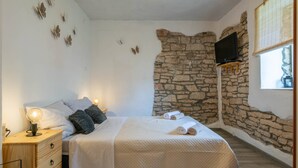 Une chambre avec un mur en pierre, un lit avec une literie blanche, une table de chevet avec une lampe et un téléviseur à écran plat fixé au mur.