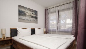 Una cama doble con ropa de cama blanca, un cabecero de madera, una mesita de noche con una lámpara y un cuadro de una escena de playa en la pared.