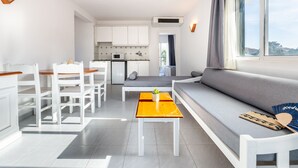 A modern living room with a sofa, dining table, and chairs, a kitchen area with cabinets and appliances, and a large window with curtains. - Vibra Caleta Playa Apartamentos (Ciutadella de Menorca)