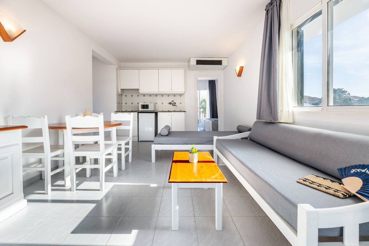 A modern living room with a sofa, dining table, and chairs, a kitchen area with cabinets and appliances, and a large window with curtains.