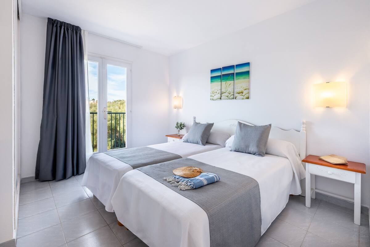 A hotel room with two beds, a balcony, a painting, and a nightstand with a lamp.