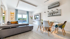 Una sala moderna con sofá, mesa de comedor y sillas. Hay un televisor montado en la pared y una ventana con cortinas.