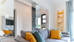 Una sala de estar moderna con un sofá gris, cojines amarillos y turquesas, un televisor montado y un estante de madera con adornos.