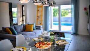 Un comedor con una mesa de vidrio, sillas y diversos alimentos. Se observa un sofá y una ventana al fondo.