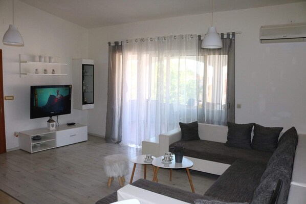 A modern living room with a flat-screen TV, white furniture, and a large window with sheer curtains.