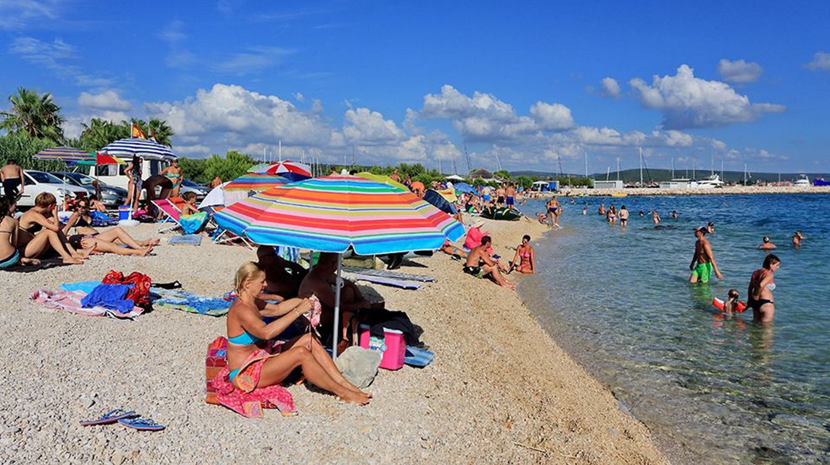 A crowded beach with people sunbathing, swimming, and under colorful umbrellas.