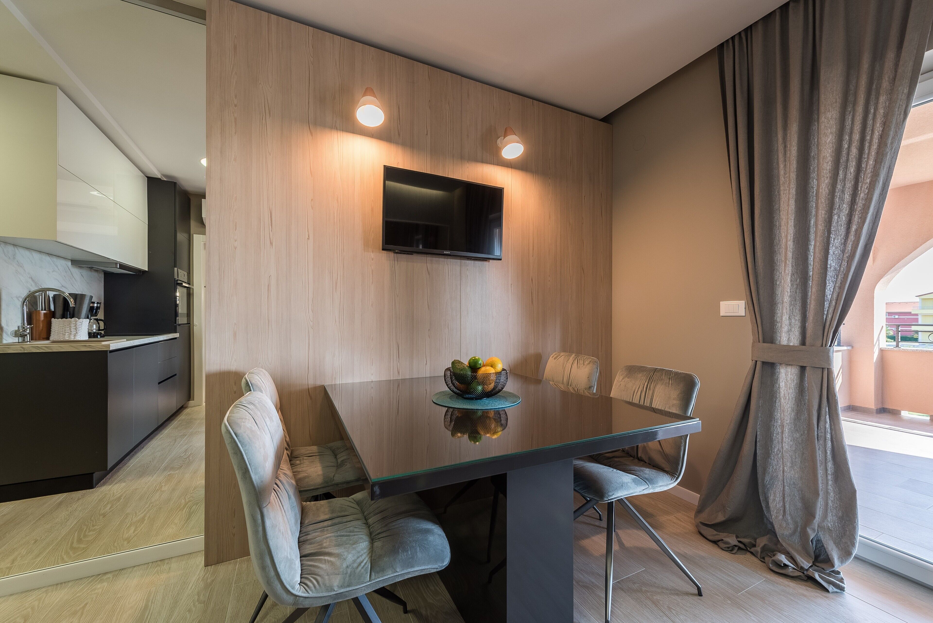 Un comedor moderno con una mesa de vidrio, seis sillas, un frutero, un televisor de pantalla plana montado en la pared y vistas al exterior a través de un ventanal grande.