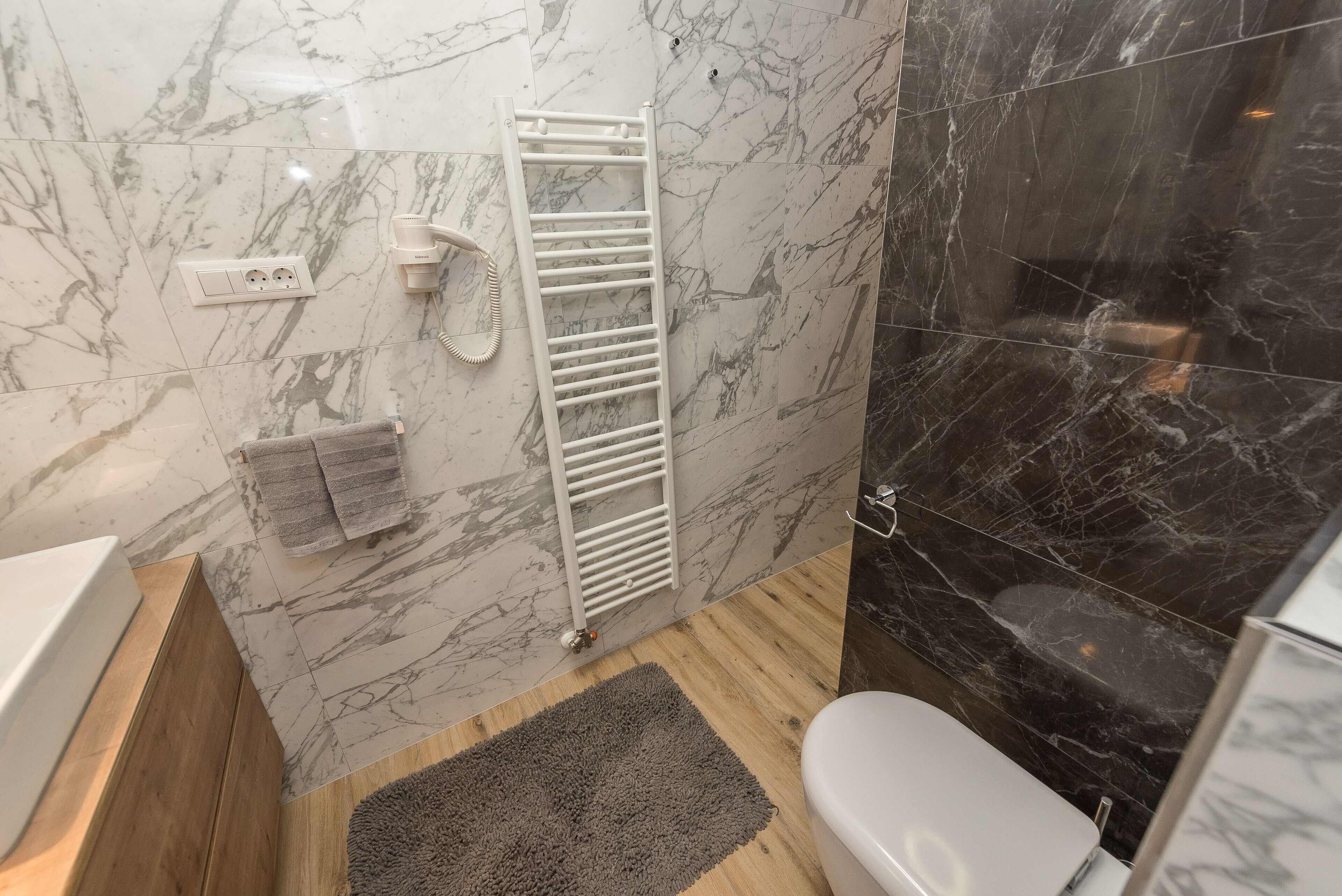 Une salle de bain avec un mur en marbre, un radiateur blanc, un porte-serviettes et un plancher en bois.