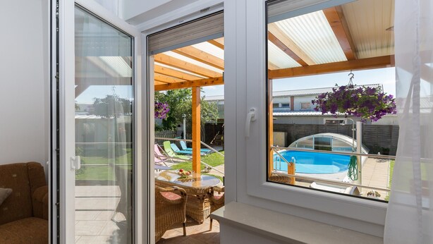 A room with a sliding glass door leading to an outdoor patio area with a pool, wicker furniture, and a hanging flower basket. - Apartment Twisting By The Pool - Two bedroom apartment with private pool (Zadar)