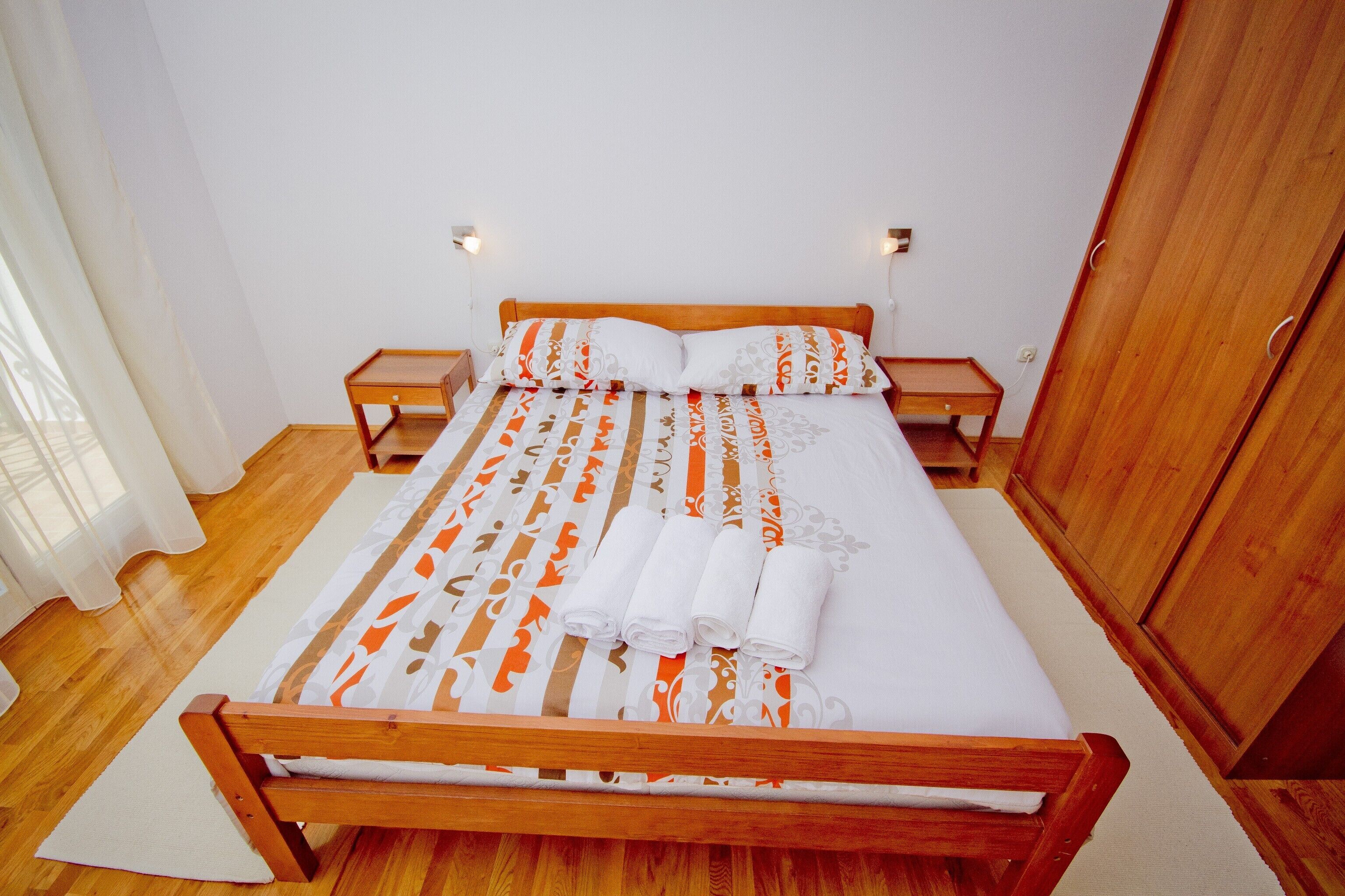 A single bed with a wooden frame, white bedding, and orange and gray patterned pillows. There are two bedside tables with lamps, a wooden wardrobe, and a window with sheer curtains.