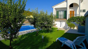 Una villa blanca con piscina, contraventanas verdes y un pequeño jardín.