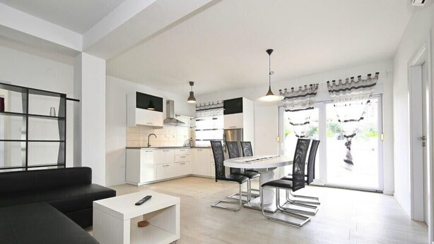 Una cocina moderna con armarios blancos, un comedor con mesa y sillas, y un sofá negro en la sala.