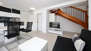 Een moderne woonkamer met een zwarte bank, een witte salontafel en zwarte netstoelen. Een televisie staat op een witte dressoir, en een houten trap is zichtbaar.