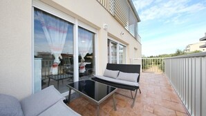 Een balkon met een glazen deur, een glazen tafel en twee grijze banken.