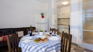 Un coin repas avec une table dressée pour deux personnes, une plante en pot et une cuisine en arrière-plan.