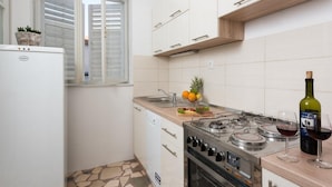 Une cuisine de taille réduite, équipée d’une cuisinière à gaz, d’un évier et d’une fenêtre avec des stores.