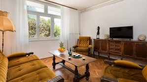 Un salon comprenant un canapé, un fauteuil et une table sur laquelle se trouvent un panier de fruits et une théière. Une télévision et des objets décoratifs sont posés sur un meuble.