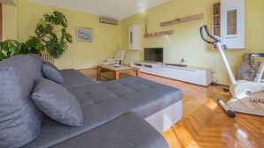 A living room with a grey sofa, a TV, a white cabinet, and a treadmill. - Apartment Villa Odesa - Two Bedroom Apartment with Balcony, shared Swimming Pool (Zadar)