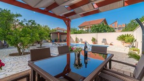 An outdoor patio with a glass-top table and wicker chairs, surrounded by a swimming pool and greenery. - Apartment Villa Odesa - Two Bedroom Apartment with Balcony, shared Swimming Pool (Zadar)