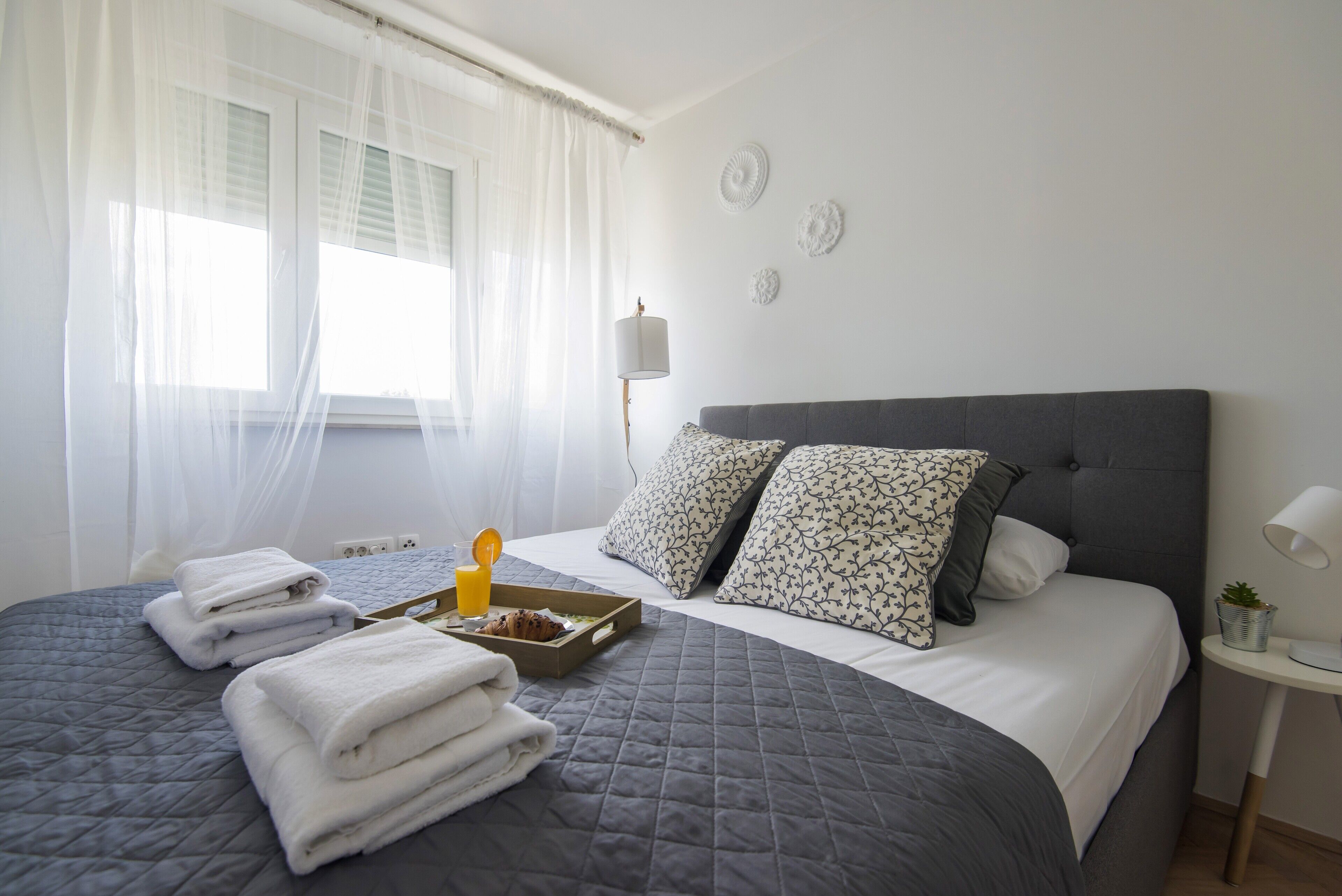 A neatly made bed with a grey quilt, white pillows, and a tray with a muffin and orange juice.