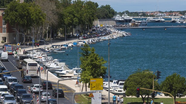 Ein belebter Hafen mit vertäuten Booten, einem Fußweg und einer Straße mit Fahrzeugen.