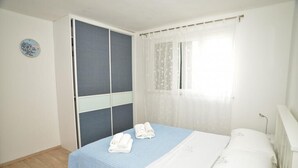 Een slaapkamer met een bed, een blauw beddegoed, witte kussens en een raam met dunne gordijnen.