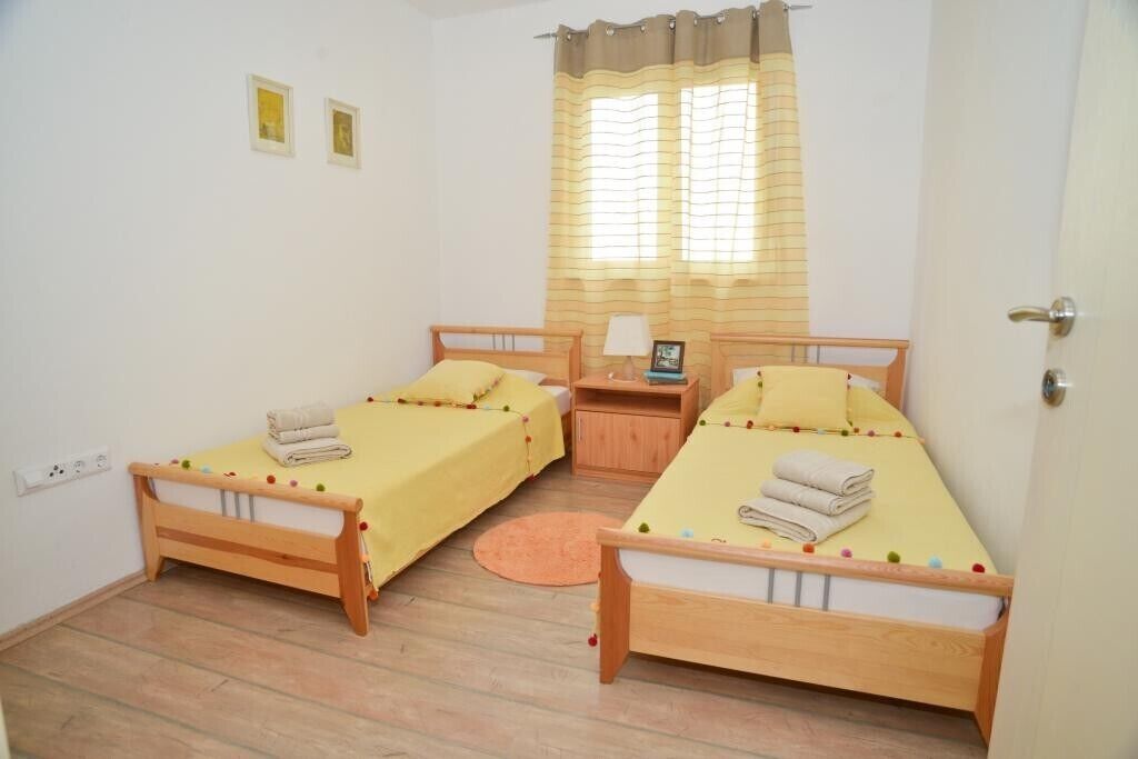 Una habitación con dos camas de madera, cada una con colcha y almohadas amarillas, una mesita de noche de madera con una lámpara, y una ventana con cortina a rayas.
