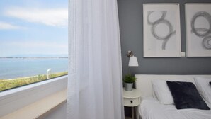 Un dormitorio con vistas al mar, una cama blanca, una mesita de noche con una lámpara y dos obras de arte abstracto en la pared.