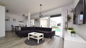 Una sala de estar moderna con un sofá modular, una mesa de centro blanca y un comedor con una mesa y sillas blancas. Una ventana grande ofrece vistas al exterior.